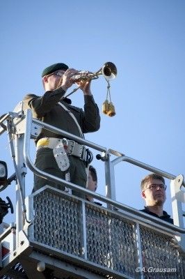 Ein Mann in Militäruniform spielt Trompete auf einer erhöhten Plattform mit klarem blauem Himmel im Hintergrund.