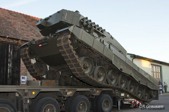 Ein Militärpanzer wird von einem Tieflader vor einem Gebäude mit Ziegeldach entladen.