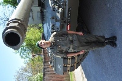 Ein Mann in Uniform steht neben einem großen Panzer mit sichtbarem Lauf und Nummernschild an der Seite.