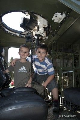 Zwei junge Jungen sitzen in einem Militärfahrzeug, einer trägt ein Adidas-Shirt und der andere ein gestreiftes Shirt. Sie posieren für ein Foto mit einer über ihnen montierten Kamera.