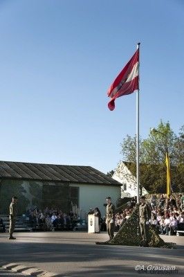Eine Flaggenhissung findet vor einem Gebäude statt, das von einer Menge Menschen beobachtet wird. Die Flagge ist rot und weiß mit einem Symbol in der Mitte.