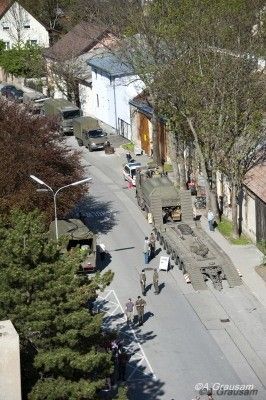Ein Luftbild einer Straße mit geparkten Militärfahrzeugen, gehenden Menschen und einem großen Militärlastwagen, der Fracht entlädt. Bäume und Gebäude sind im Hintergrund sichtbar.