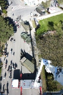 Eine Luftaufnahme einer Straße mit Menschen, die gehen, und einem Feuerwehrwagen und Militärfahrzeugen, die am Straßenrand geparkt sind. Bäume und Gras befinden sich am Straßenrand. Ein Wasserzeichen befindet sich unten rechts.