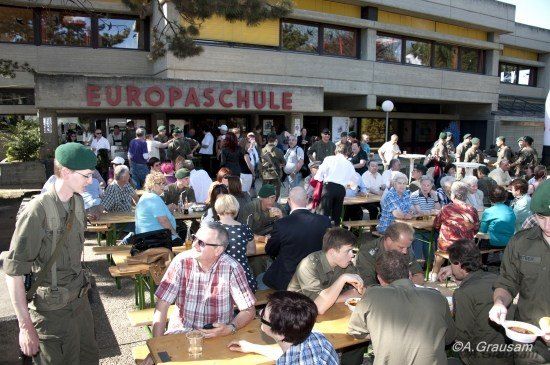 Eine Gruppe von Menschen in Militäruniformen und Zivilkleidung essen in einem Outdoor-Restaurant vor einem Gebäude namens 'EUROPASCHULE'.