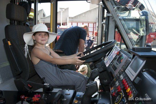 Ein junges Mädchen mit einem breitkrempigen Hut sitzt auf dem Fahrersitz eines Feuerwehrwagens, während ein Mann hinter ihr steht und aus dem Fenster schaut.