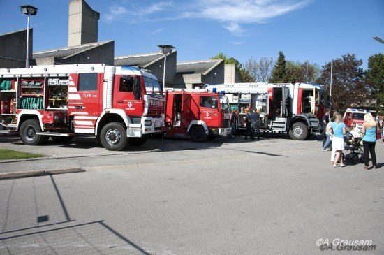 Drei rote Feuerwehrfahrzeuge sind auf einem Parkplatz geparkt, um die herum Menschen stehen. Die Fahrzeuge sind mit verschiedenen Werkzeugen und Ausrüstung ausgestattet.