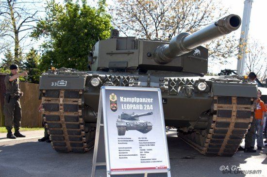 Ein großer Militärpanzer, gekennzeichnet als Kampfpanzer Leopard 2A4, ist im Freien ausgestellt. Er hat eine große Kanone und ist von Menschen und Bäumen umgeben.