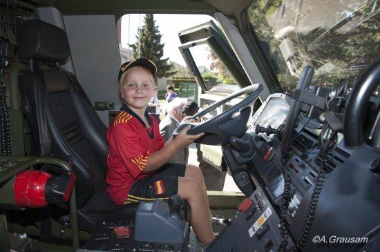 Ein kleiner Junge in einem roten Shirt und Hut sitzt im Fahrersitz eines Militärfahrzeugs und lächelt.