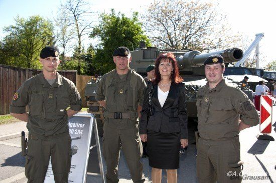 Drei Soldaten in Militäruniformen stehen neben einer Frau in einem Anzug vor einem Panzer.