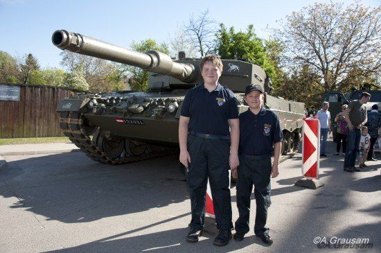 Zwei Männer in Uniform stehen lächelnd vor einem Militärpanzer, mit einer Menge und einem Holzzaun im Hintergrund.