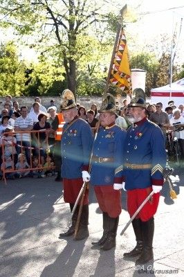 Drei Männer in blauen Uniformen und roten Hosen stehen bei einer Parade, halten Fahnen und Gewehre, während eine Menge im Hintergrund zuschaut.