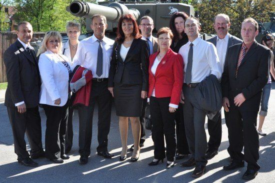 Eine Gruppe formell gekleideter Personen posiert für ein Foto vor einem Militärfahrzeug.