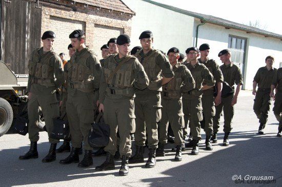 Eine Gruppe von Soldaten in grünen Uniformen, mit Mützen und Stiefeln, marschiert in einer Reihe vor einem Gebäude.