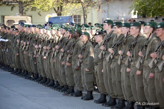 Eine Reihe von Soldaten in grünen Uniformen und Mützen steht in Formation, Gewehre haltend, mit Gebäuden und Bäumen im Hintergrund.
