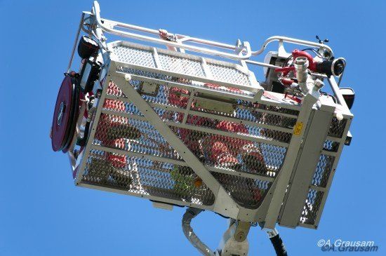 Ein Feuerwehrmann in roter Uniform befindet sich in einem Metallkorb, der an einem Feuerwehrauto befestigt ist, möglicherweise auf einer Rettungsmission.