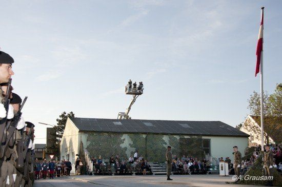 Eine militärische Zeremonie findet vor einem Gebäude mit Tarndesign statt. Auf der rechten Seite ist eine Flagge auf einem Mast gehisst.