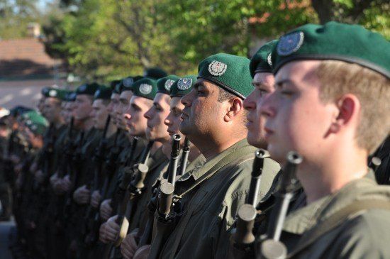Eine Reihe von Soldaten in grünen Uniformen und Baretten steht in Formation und hält Gewehre. Sie schauen nach vorne, möglicherweise während einer Parade.