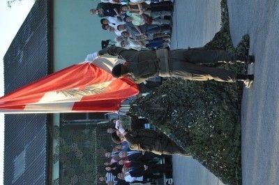 Ein Militäroffizier hält eine Flagge, während eine andere Person vor ihm kniet. Eine Menschenmenge steht hinter ihnen und beobachtet die Veranstaltung.