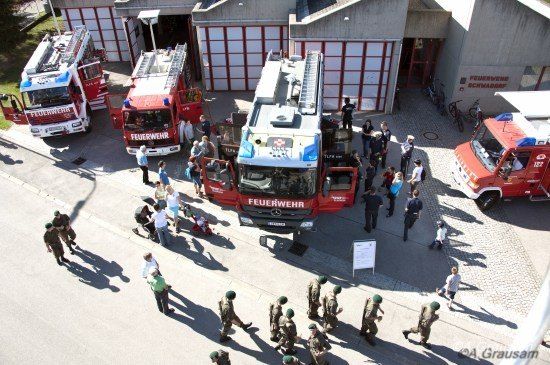 Eine Vogelperspektive auf eine Feuerwache mit mehreren Feuerwehrfahrzeugen, die draußen geparkt sind. Feuerwehrleute und Zivilisten sind um die Fahrzeuge versammelt.
