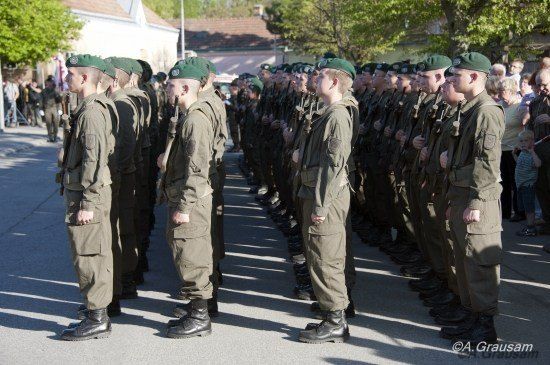 Eine Reihe von Soldaten in grünen Uniformen steht in Formation auf einer Straße, mit Zuschauern dahinter.