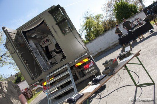 Ein Militärfahrzeug mit geöffneter Hecktür, das Innenausstattung zeigt. Zwei Soldaten stehen in der Nähe, einer hält eine Waffe, vor ihnen steht ein Tisch und Ausrüstung.