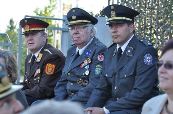 Drei Männer in Militäruniformen sitzen zusammen, einer mit Brille, ein anderer mit einem roten Kreuz auf der Brust und der dritte mit einem roten Abzeichen auf der Schulter. Sie nehmen wahrscheinlich an einer formellen Veranstaltung teil.