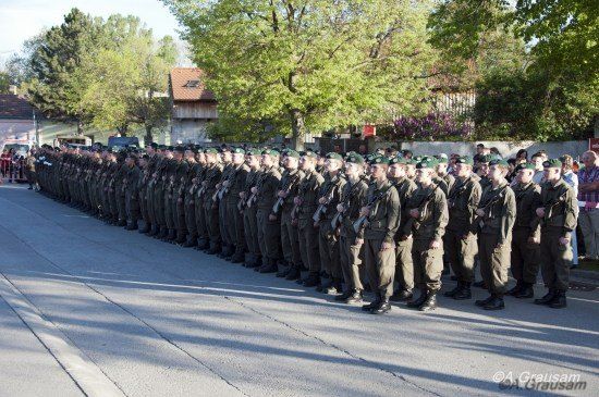 Eine Reihe von Soldaten in Uniform, Gewehre haltend, in Formation auf einer Straße stehend, mit Bäumen und Gebäuden im Hintergrund.