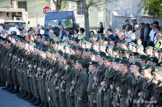 Eine große Gruppe von Militärangehörigen in grünen Baretten und Uniformen steht in einer Reihe und hält Gewehre, während eine Menschenmenge im Hintergrund zuschaut.