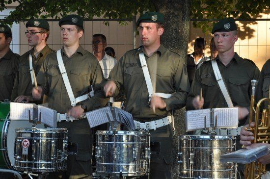 Fünf Männer in Militäruniformen spielen Trommeln und Trompeten im Freien. Sie haben Notenständer mit Notenblättern vor sich.