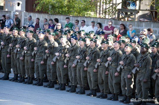 Eine Reihe von Soldaten in grünen Uniformen und Mützen steht in Formation, Gewehre in der Hand, mit einer Menge von Zuschauern hinter ihnen.