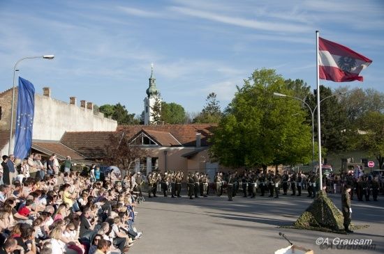 Ein Militärorchester spielt an einem sonnigen Tag vor einer Menge, mit einem Fahnenmast und einer Kirche im Hintergrund.