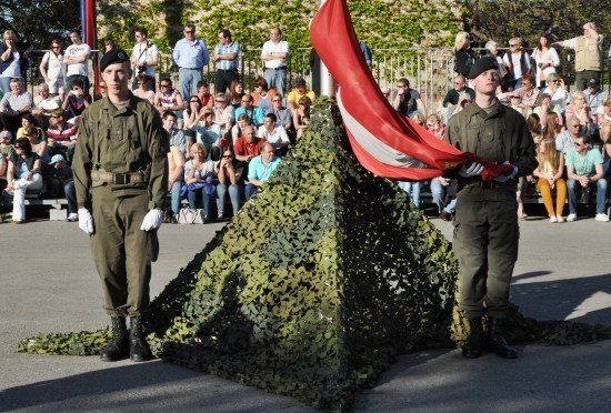 Zwei Soldaten stehen neben einer getarnten Pyramide und halten eine rote Flagge, während eine Menge hinter einem Zaun zusieht.