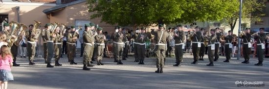 Eine Militärkapelle in Uniform steht in Formation auf einer Straße, eine junge Mädchen in einem rosa Shirt beobachtet von der Seite.