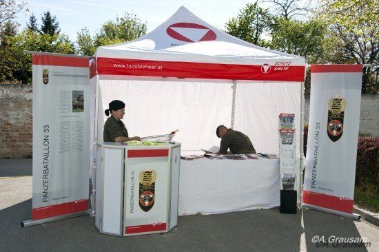 Zwei Personen arbeiten an einem Stand unter einem weißen und roten Zelt, mit einer Backsteinmauer und Bäumen im Hintergrund. Das Zelt hat Banner mit dem Text 'Panzerbataillon 33' und einem Logo.