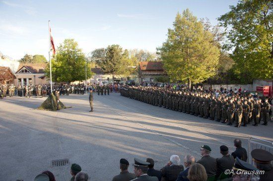 Ein Militärparade mit Soldaten in Uniform, die in Reihen stehen, einem Fahnenmast im Hintergrund und Zuschauern, die von den Seiten zuschauen.