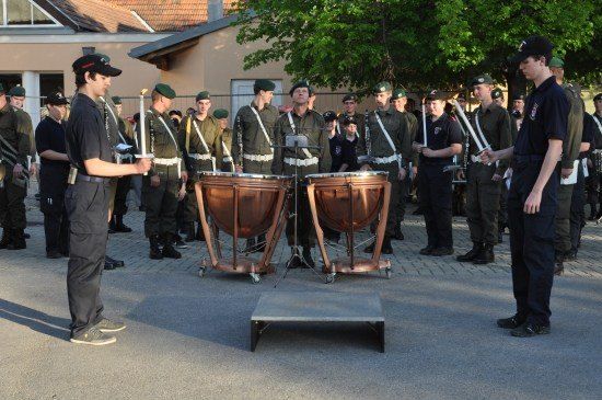 Eine Gruppe von Militärangehörigen in Uniform steht in Formation, wobei zwei Personen Trommeln spielen und andere Fackeln halten. Sie stehen vor einem Gebäude mit einem Baum.