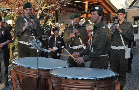Eine Militärkapelle spielt im Freien, wobei zwei Schlagzeuger große Trommeln schlagen, während andere Bandmitglieder Klarinetten und Trompeten spielen.