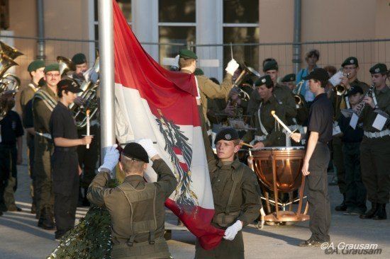 Militärangehörige in Uniform hissen eine Flagge mit Wappen, während eine Band im Hintergrund spielt.
