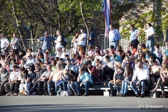 Eine Menschenmenge sitzt auf Tribünen, einige mit Sonnenbrillen, andere mit Hüten, und beobachtet eine Veranstaltung. Bäume und eine Fahne sind im Hintergrund zu sehen.