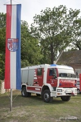 Ein weißer und roter Feuerwehrwagen steht auf dem Gras mit einem Fahnenmast davor. Die Fahne hat einen Löwen und ein Wappen darauf.