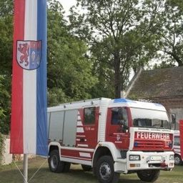 Ein weißer und roter Feuerwehrwagen steht auf dem Gras mit einem Fahnenmast davor. Die Fahne hat einen Löwen und ein Wappen darauf.