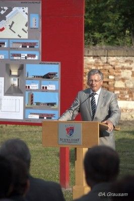 Ein Mann in Anzug und Krawatte spricht an einem Podium vor einem großen Poster, mit mehreren Menschen im Hintergrund.