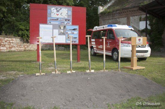 Ein roter Schilderkasten mit Diagrammen steht neben einem Haufen Erde mit Schaufeln, mit einem geparkten Feuerwehrwagen in der Nähe.