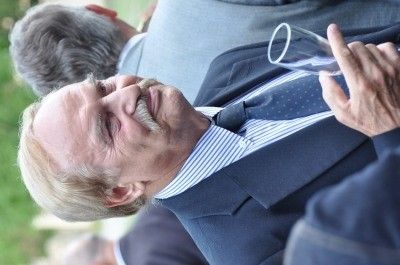 Ein älterer Mann mit Schnurrbart und blauem gestreiftem Hemd schaut nach oben und hält ein Weinglas.