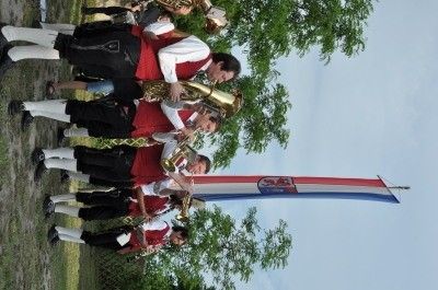 Eine Gruppe von Musikern in traditioneller Kleidung spielt Blechblasinstrumente im Freien, mit einer kroatischen Flagge im Hintergrund.