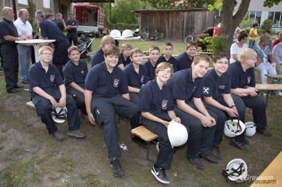 Eine Gruppe junger Männer in Uniformen und Helmen sitzt draußen auf Bänken, mit Erwachsenen im Hintergrund, die stehen und sitzen.