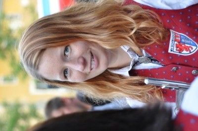Eine Frau mit langen blonden Haaren und blauen Augen, die eine rote gepunktete Weste mit einem Logo trägt, lächelt und in die Kamera schaut.