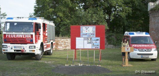 Zwei Feuerwehrfahrzeuge und ein Krankenwagen stehen auf einem Rasenfeld. Eine Frau steht an einem Podium vor einem roten Schild mit Text und Bildern. Bäume und eine Steinmauer sind im Hintergrund.