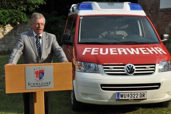Ein Mann in einem Anzug steht an einem Podium neben einem roten und weißen Feuerwehrauto mit dem Wort 'Feuerwehr' darauf.