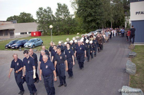Eine Gruppe Feuerwehrleute in Uniform, einige mit weißen Helmen, gehen eine Straße entlang, an der geparkte Autos und Bäume stehen. Im Hintergrund ist ein Gebäude mit den Buchstaben 'PFA' zu sehen.